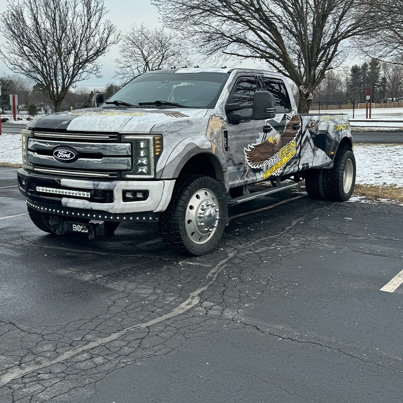 A customized Ford pickup truck with a 'Top Gen Roofing' wrap featuring an eagle graphic, parked on a snowy asphalt lot with trees in the background.